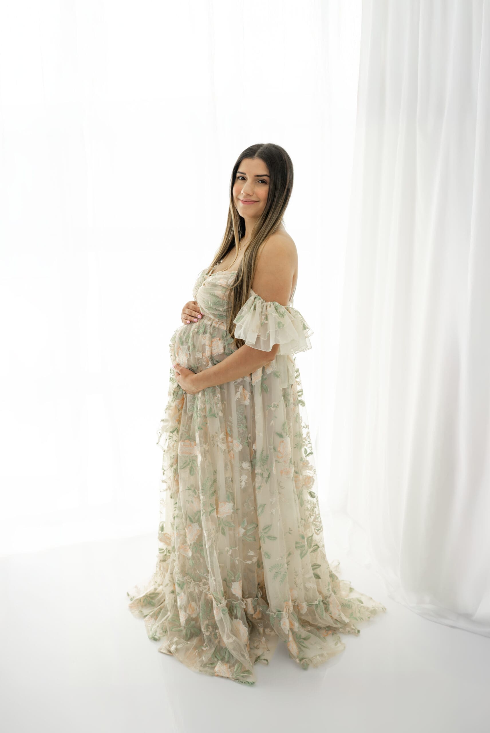 Pregnant woman in floral maternity gown standing in front of window smiling — captured by Alliston and Barrie maternity, newborn, and family photographer.